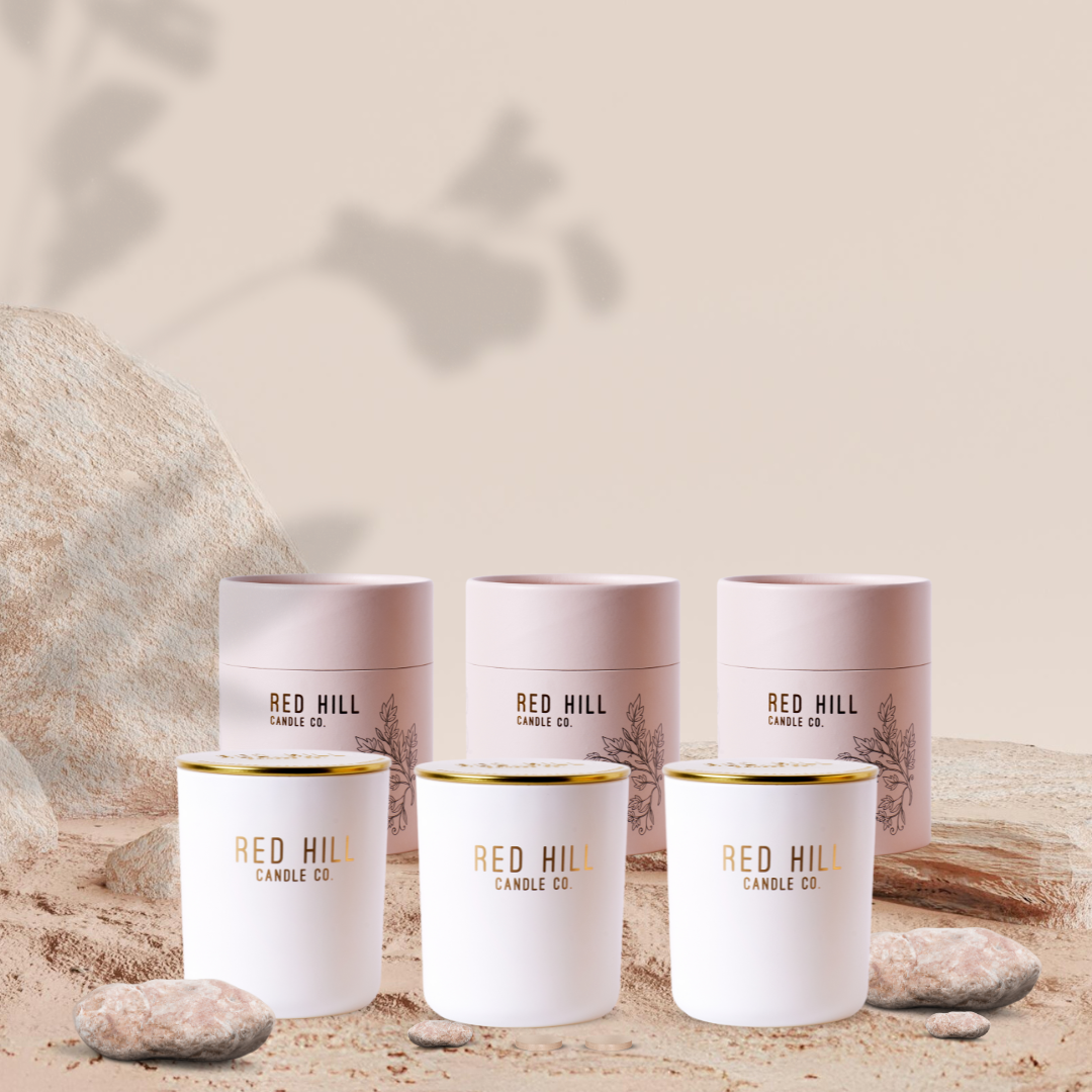 Three large candles from Red Hill Candle Co., displayed in white containers with gold accents, set against a neutral background with rocks and a shadow of a plant.