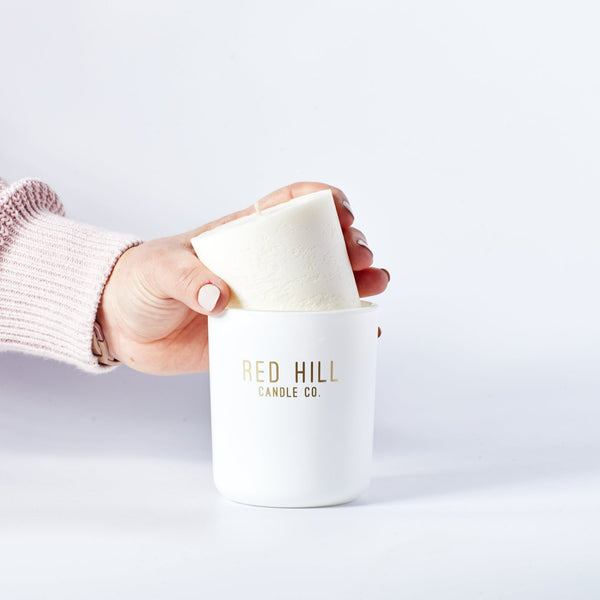 A hand lifting a white candle refill with the Red Hill Candle Co. branding on it